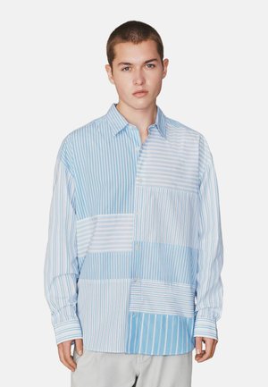 Mladý muž, ktorý nosí modro-bielu pruhovanú košeľu so vzorom z nášiviek a dlhými rukávmi, stojaci pred jednoduchým pozadím.