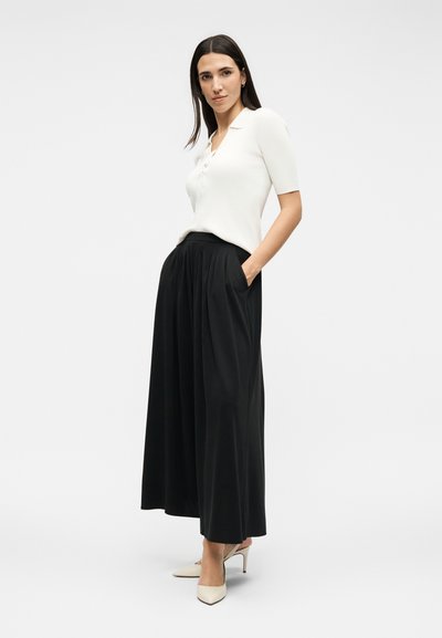 Γυναίκα στέκεται με τα χέρια στις τσέπες, φοράει λευκό κοντομάνικο πόλο, μαύρο φαρδύ παντελόνι και μπεζ ψηλοτάκουνες γόβες.