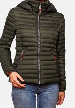 Kvinde iført en olivengrøn quiltet jakke med lynlås og to lynlåslommer samt blå jeans, stående mod en ensfarvet baggrund.