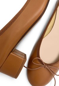 Ballerine in pelle marrone con tacco quadrato. Le caratteristiche includono una punta rotonda, bordi cuciti e un fiocco decorativo sulla parte anteriore.
