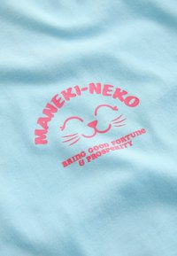 Svetlo modra bombažna majica z rožnato grafiko nasmejanega mačka ter napisom "MANEKI-NEKO" in "PRINAŠA DOBRO SREČO IN BLAGOSTANJE."