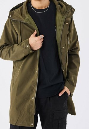 Personne portant une veste à capuche vert olive par-dessus une chemise noire et un pantalon cargo noir, tenant la fermeture éclair de la veste d'une main.