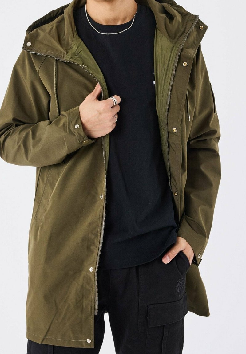Personne portant une veste à capuche vert olive par-dessus une chemise noire et un pantalon cargo noir, tenant la fermeture éclair de la veste d'une main.