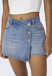 Vaaleansininen farkkuhame, jossa on napillinen edestä, rispaantunut helma ja kaksi etutaskua. Materiaali on haalistuneen näköinen.