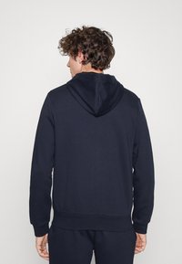 Hanorac de culoare albastru marin, cu glugă, având o textură netedă, mâneci lungi și un tiv elastic. Design simplu, fără logo-uri sau modele vizibile.