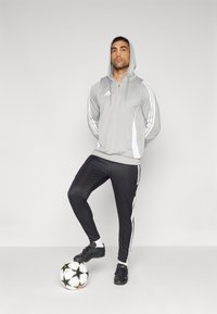 Grijze hoodie met rits en witte accenten en een drievoudig streepdesign, gecombineerd met zwarte sportbroeken met zijstrepen. Zwarte voetbalschoenen, voetbal.