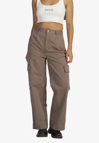 Baggy cargo pants in brown cotton with side pockets, a flat front, and a high waist design. Paired with a white cropped tank top.