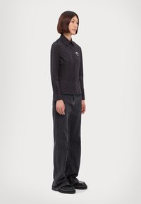 Donna con capelli a caschetto lisci indossa una camicia nera con bottoni, jeans scuri larghi e scarpe platform nere, in piedi contro uno sfondo bianco semplice.