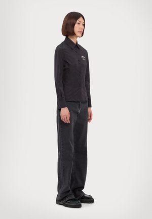 Schwarzes langärmliges Hemd mit Knöpfen und Kragen, mit einem dezenten Logo, kombiniert mit locker sitzenden dunklen Jeans und schwarzen Plateauschuhen.