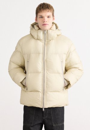 Beige puffervinterjakke med høj krave, to lynlåslommer i siden og en glat, quiltet tekstur, der har en fuld frontlynlås.