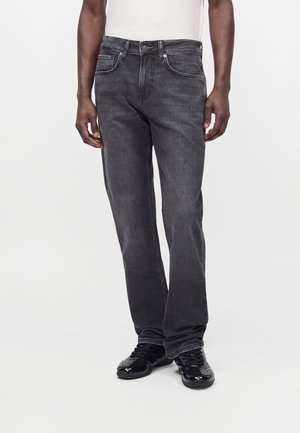 Homme portant un jean noir délavé à jambes droites et des baskets noires, debout sur un fond blanc.