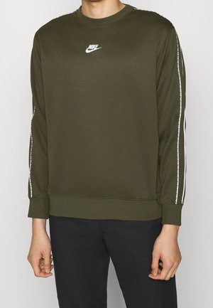 Persona che indossa una felpa a maniche lunghe Nike verde oliva con logo bianco sul petto e dettagli a strisce bianche sulle maniche, abbinata a pantaloni neri.