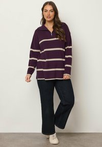 Lila und beige gestreifte Langarmbluse mit Halbreißverschluss-Kragen und Seitenschlitzen, kombiniert mit dunklen Jeans und hellen Sneakern.