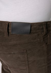 Bruine corduroy broek met een gestructureerde afwerking, gestikte details en een zwart leren label op de tailleband met de tekst "HEREN+".