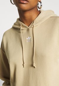 Sudadera con capucha beige con cordón, hecha de tela suave, con un logo de Adidas bordado en blanco en el pecho. Textura suave y ajuste relajado.