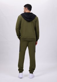 Olivgrüner Trainingsanzug mit schwarzer Kapuze, gerippten Bündchen, Seitentaschen und schmal zulaufenden Hosen. Das Material wirkt weich und strukturiert. Weiße Sneakers.