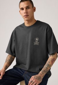 Άνδρας με τατουάζ στα χέρια φοράει μια φαρδιά σκούρα γκρι μπλούζα με ένα μικρό σχέδιο λουλουδιού και κείμενο στο στήθος, καθισμένος μπροστά σε απλό φόντο.