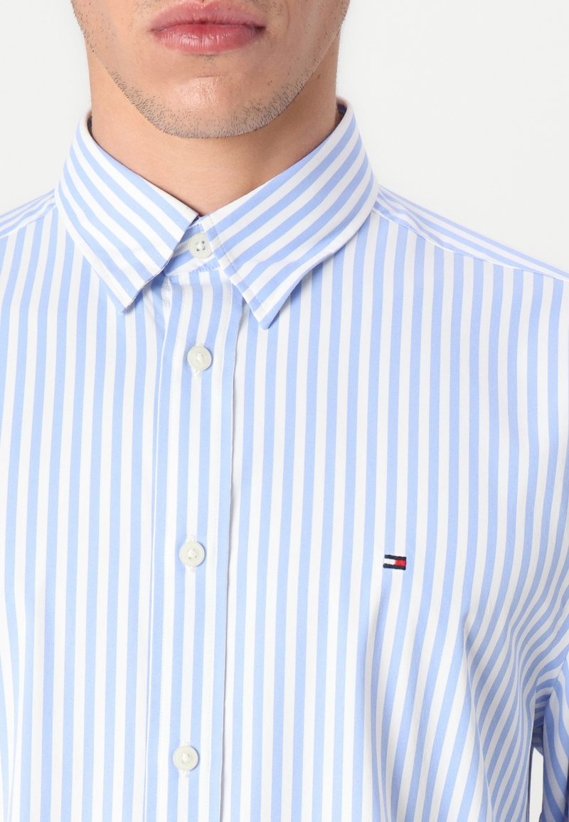 Chemise à rayures bleu clair et blanche ; dotée d'un col et d'une patte de boutonnage, avec un petit logo sur la poitrine.