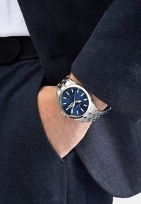 Silberner Edelstahlmann mit blauer Zifferblatt, leuchtenden Markierungen und Datumsanzeige, getragen an einem Handgelenk mit einem navyblauen strukturierte Stoffblazer.