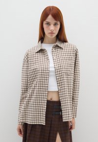 Camicia a quadri marrone e beige con colletto classico, vestibilità rilassata e taschino singolo sul petto, indossata sopra una maglietta corta bianca.