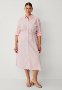 Lichtroze knopenjurk gemaakt van zachte stof. Kenmerkt een elastische taille, opgerolde mouwen en een lengte tot halverwege de kuit. Gekleed met beige blokhak schoenen.