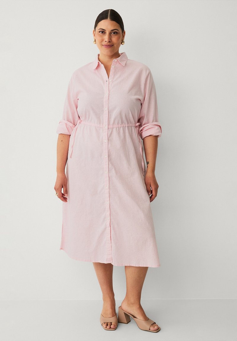 Lichtroze knopenjurk gemaakt van zachte stof. Kenmerkt een elastische taille, opgerolde mouwen en een lengte tot halverwege de kuit. Gekleed met beige blokhak schoenen.