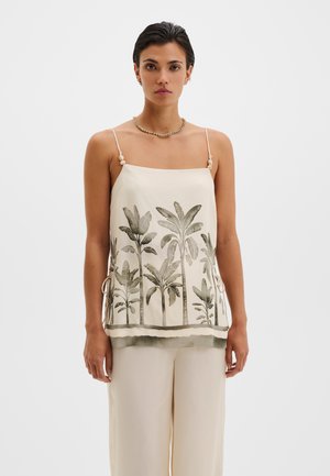 Femme portant un caraco crème à fines bretelles avec un imprimé de palmier, associé à un pantalon ample crème, debout devant un fond blanc.