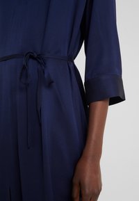 Bras à la peau foncée à côté d'une robe texturée bleu marine avec des manches trois-quarts et une ceinture nouée à la taille.