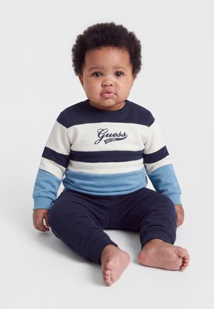 Bébé aux cheveux bouclés, assis pieds nus, portant un sweat-shirt Guess rayé bleu, blanc et noir et un pantalon bleu foncé sur fond blanc.