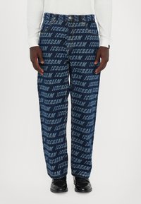 Denim pants in dark blue with repeated "ICECREAM" text pattern in lighter blue. Loose fit with standard pockets, worn with black shoes.