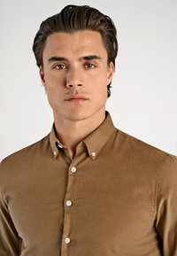 Camicia marrone con bottoni, realizzata in un tessuto liscio, con bottoni bianchi e colletto appuntito. Il design è slim fit e a maniche lunghe.