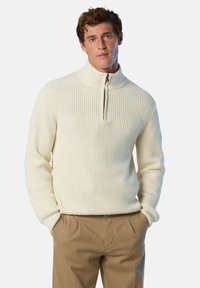 Maglione a girocollo con zip a un quarto in maglia cremosa, con texture a coste e colletto alto, abbinato a pantaloni marrone chiaro, mani in tasca.