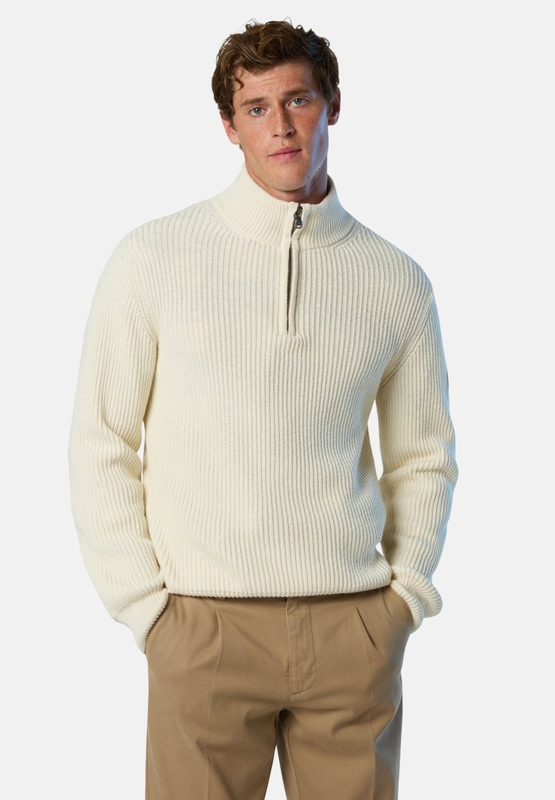 Maglione a girocollo con zip a un quarto in maglia cremosa, con texture a coste e colletto alto, abbinato a pantaloni marrone chiaro, mani in tasca.