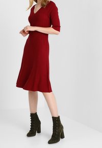 Abito a maglia rosso a costine con scollo a V e maniche corte, svasato all'orlo. Abbinato a stivaletti alla caviglia in suede verde scuro con lacci e bottoni.