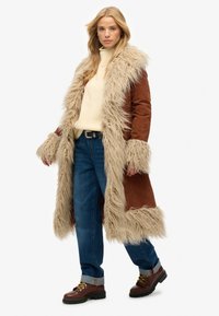 Brun faux fur-foret coat med en beige shearling krave, parret med en cremefarvet sweater, blå jeans og brune snørebåndsstøvler.