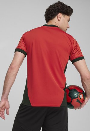 Rood en groen sportshirt met een gestructureerd patroon op de mouwen, korte mouwen en een hoge ronde hals; zwarte shorts; houdt een gekleurde voetbal vast.