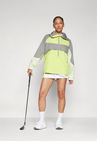 Lightweight, two-tone windbreaker with a gray and neon green design. Features a half-zip closure, hood, and elasticized cuffs. Positioned with a golf club.