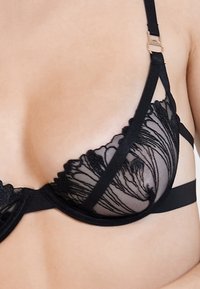 Bralette de renda preta com copas transparentes e bordados florais, alças ajustáveis e uma faixa de suporte lisa. Os detalhes intrincados realçam a textura.