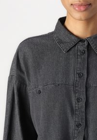 Camisa botânica cinza escura com colarinho, apresentando um tecido ligeiramente texturizado, bolsos frontais e botões pretos.