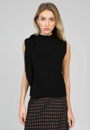 Femme aux longs cheveux blonds portant un haut noir sans manches en maille et une jupe noire à pois marron sur un fond gris.