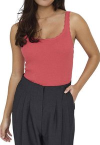 Top côtelé sans manches en corail avec un col festonné, porté avec un pantalon taille haute gris comportant des plis et des poches latérales.