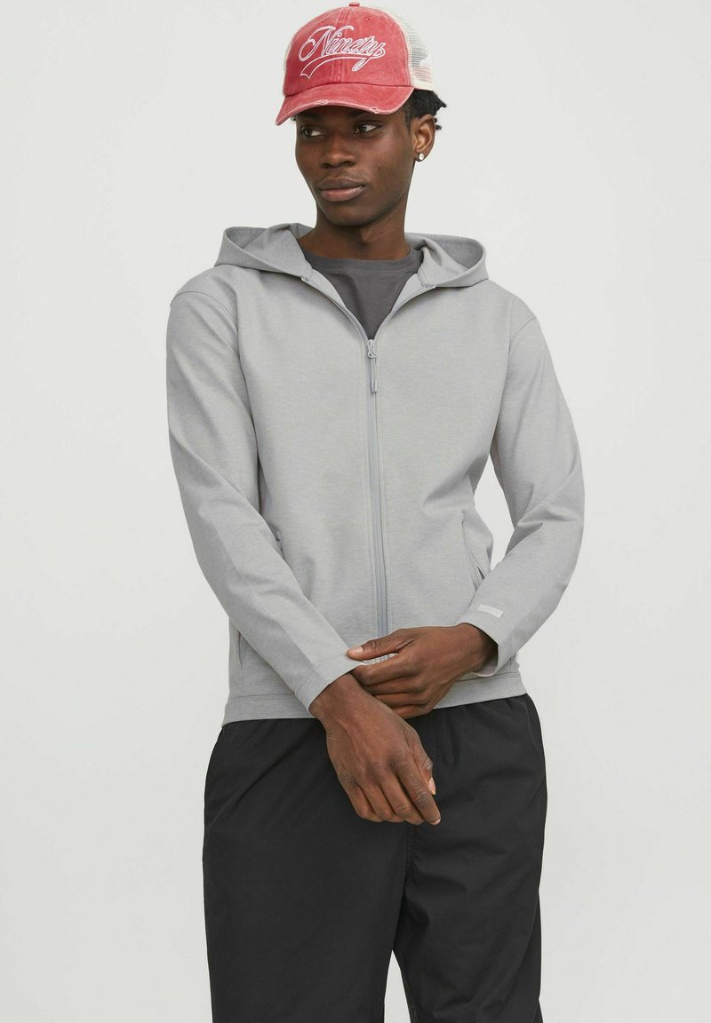Sudadera de cremallera de color gris claro con capucha, mangas largas y bolsillos laterales. Textura suave, combinada con pantalones negros y una gorra de béisbol roja.