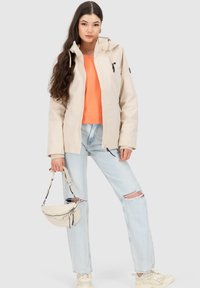 Giacca impermeabile beige con cappuccio, zip frontale e dettagli neri, indossata sopra una maglietta arancione e jeans strappati azzurri chiari. Borsa bianca e sneakers.