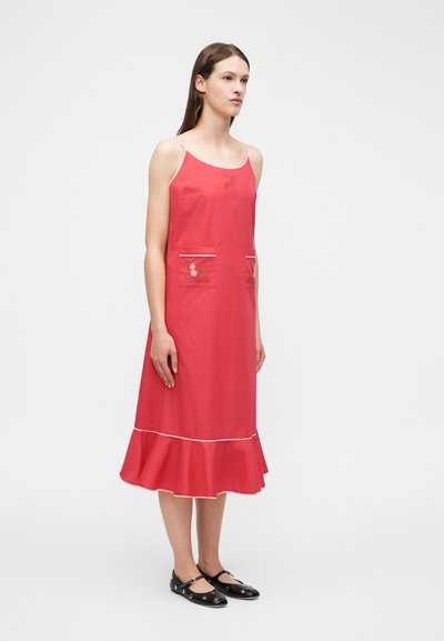 Γυναίκα που φοράει αμάνικο κόκκινο μεσοφόρι με κεντήματα λουλουδιών, τσέπες στο μπροστινό μέρος, βολάν στο κάτω μέρος και μαύρα παπούτσια τύπου Mary Jane.