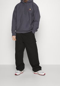 Mężczyzna w ciemnoszarej bluzie Carhartt, luźnych czarnych spodniach i białych sneakersach z czerwonymi podeszwami, stojący na tle jednolitego białego tła.
