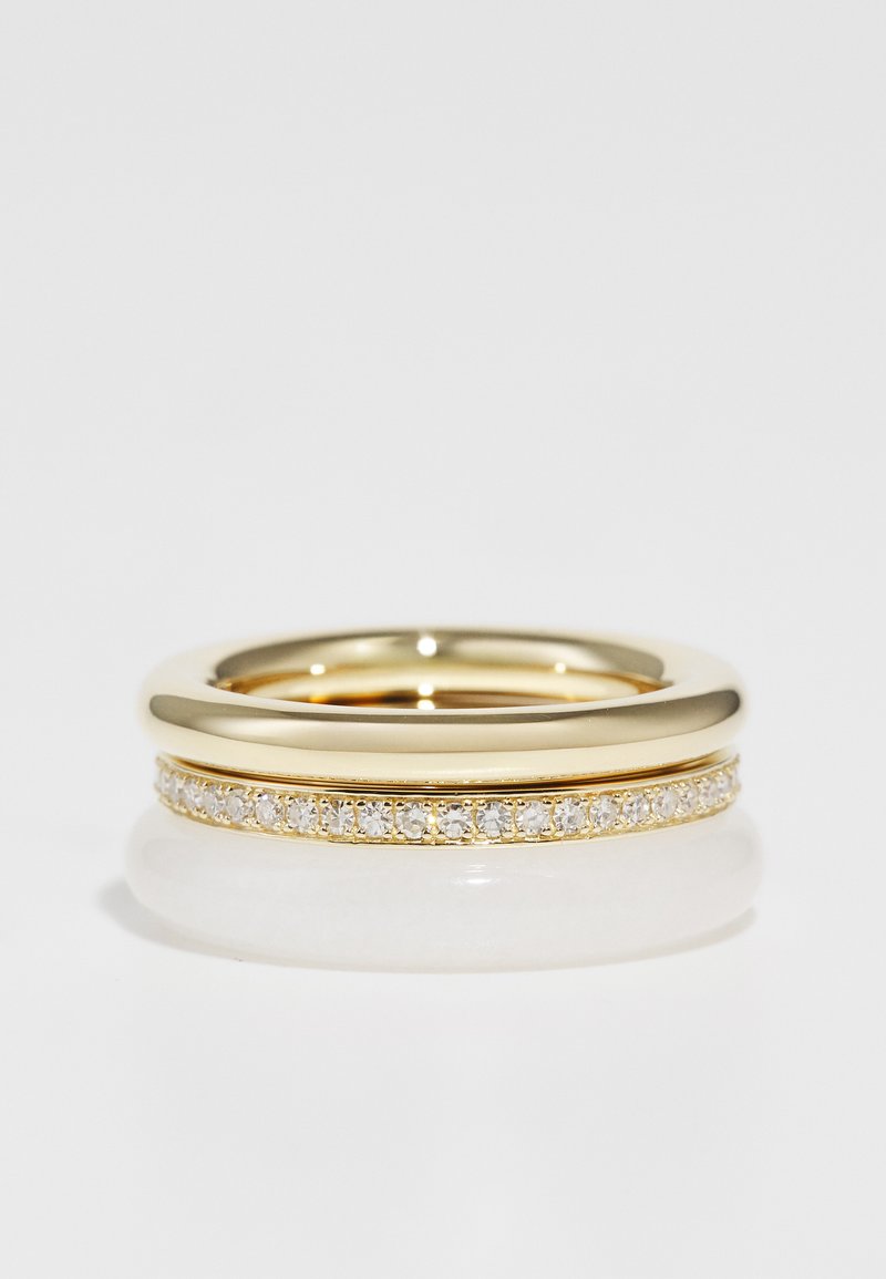 Guldring set med en slät, bred ring, överlagd av en smal ring prydd med små klara stenar. Cirkulär form, polerad yta.