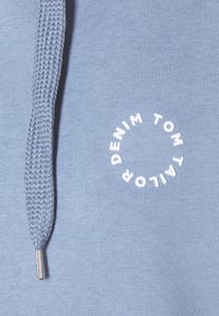 Ljusblå sweatshirt med strukturerad dragsko. Har en rund vit logotyp som tydligt står "TOM TAILOR DENIM".