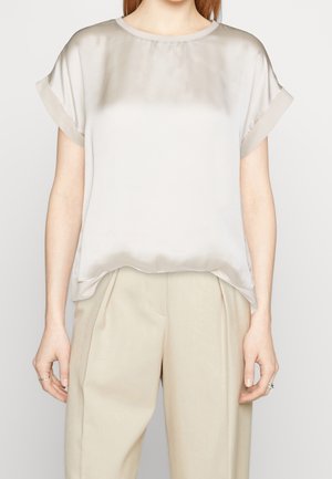 Femme portant un chemisier ample en satin beige clair à manches courtes, rentré dans un pantalon crème plissé taille haute, debout, les bras détendus.