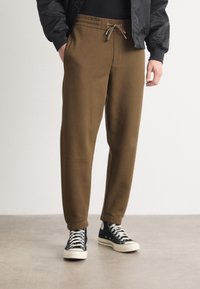 Braune Sweatpants mit elastischem Bund und Kordelzug, ausgestattet mit Seitentaschen und schmal zulaufenden Bündchen, kombiniert mit schwarzen High-Top-Sneakers.