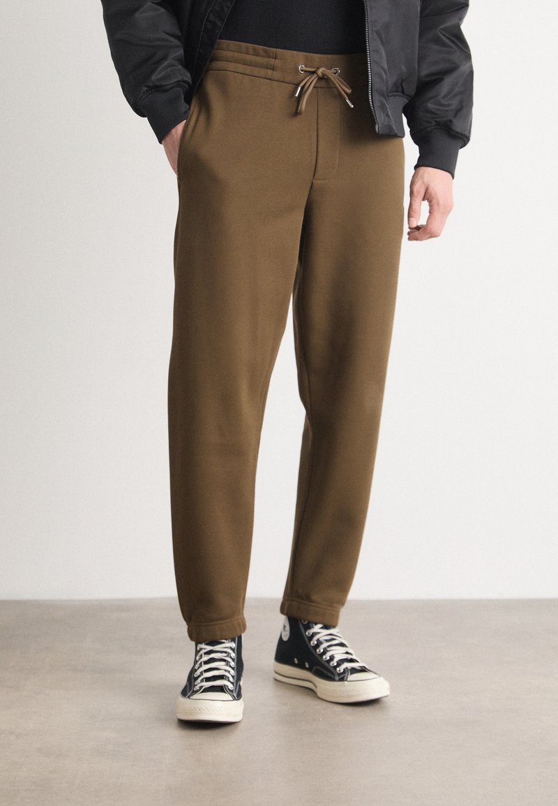 Braune Sweatpants mit elastischem Bund und Kordelzug, ausgestattet mit Seitentaschen und schmal zulaufenden Bündchen, kombiniert mit schwarzen High-Top-Sneakers.
